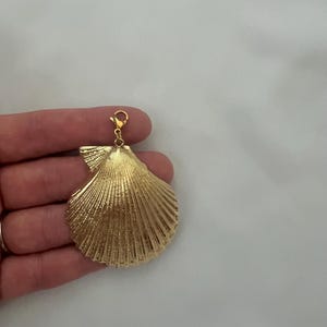 May include: A gold-toned metal seashell charm featuring a textured, ribbed surface and a small lobster claw clasp attached to the top. The shell has a fan-like shape with distinct radiating lines extending from the hinge to the scalloped edge.