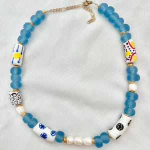 Könnte beinhalten: Handgefertigte Halskette mit hellblauen Glasperlen, weißen zylindrischen Perlen mit bunten Mustern und Süßwasserperlen. Die Halskette hat eine goldfarbene Kette und einen Verschluss, was ein einzigartiges Accessoire ergibt.