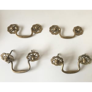 Op de afbeelding: Set van zes antieke messing lade grepen. Elke greep heeft een bloemenontwerp op de achterplaat en een gebogen handvat. De grepen zijn warm messingkleurig en zijn gerangschikt op een wit oppervlak.