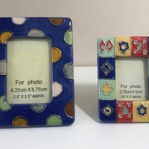 Könnte beinhalten: Zwei dekorative Keramik-Fotorahmen. Der größere Rahmen ist dunkelblau mit bunten Tupfen. Der kleinere Rahmen ist mit geometrischen Formen in Rot, Gelb, Blau und Grün verziert. Der Text auf den Rahmen lautet "Für Foto" und die Abmessungen.
