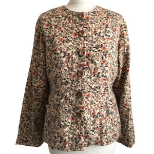 May include: A long-sleeved jacket featuring a cream base with a dense floral pattern in shades of coral, muted blue, and dark brown. The jacket has a round neckline, five large brown buttons down the centre front, and two integrated front pockets.