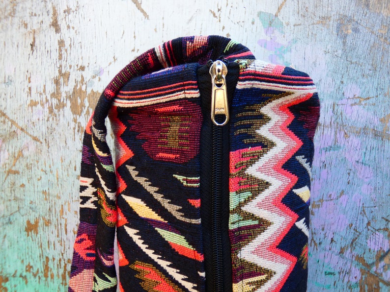 May include: A black yoga mat bag with a zipper closure. The bag is made of a colorful fabric with a geometric pattern in shades of red, pink, white, yellow, green, and blue.