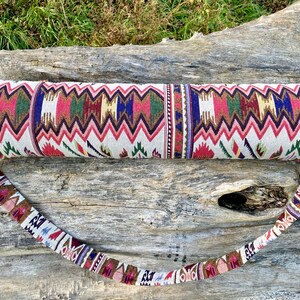 Pu&ograve; includere: Una borsa per tappetino da yoga fantasia con cinturino abbinato. La borsa presenta un motivo a zig-zag ripetuto in rosso, rosa, verde, blu e oro su sfondo bianco. Il cinturino ha un motivo simile. La borsa &egrave; cilindrica.
