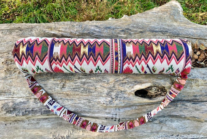 Pu&ograve; includere: Una borsa per tappetino da yoga fantasia con cinturino abbinato. La borsa presenta un motivo a zig-zag ripetuto in rosso, rosa, verde, blu e oro su sfondo bianco. Il cinturino ha un motivo simile. La borsa &egrave; cilindrica.