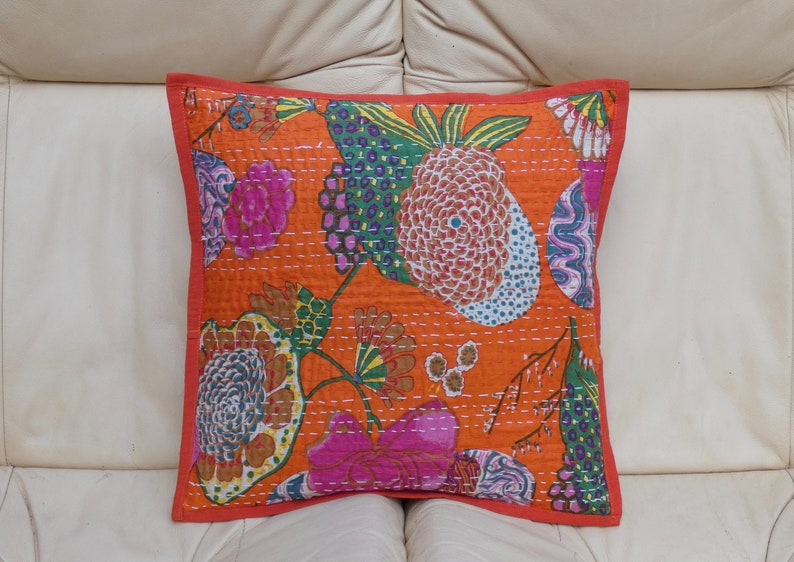 Peut inclure: Coussin carr&eacute; orange avec un motif floral rose, vert, bleu et blanc. Le coussin a une bordure rouge et des coutures blanches. Le coussin est pos&eacute; sur une surface blanche.