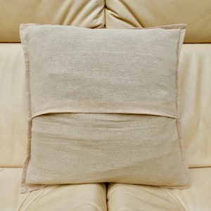Puede incluir: Una almohada rectangular lisa de color blanco roto con una cremallera en la parte posterior. La almohada est&aacute; hecha de una tela suave y natural y est&aacute; sentada en un sof&aacute; de cuero marr&oacute;n claro.