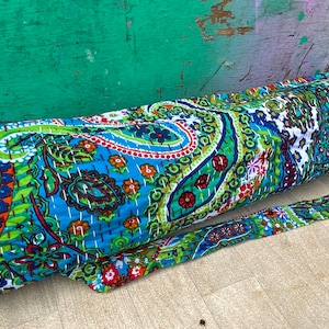 May include: A patterned yoga mat bag in shades of blue, green, orange, and white. The bag features a paisley and floral design and has a long strap for carrying. The bag is made of quilted fabric and is cylindrical in shape.
