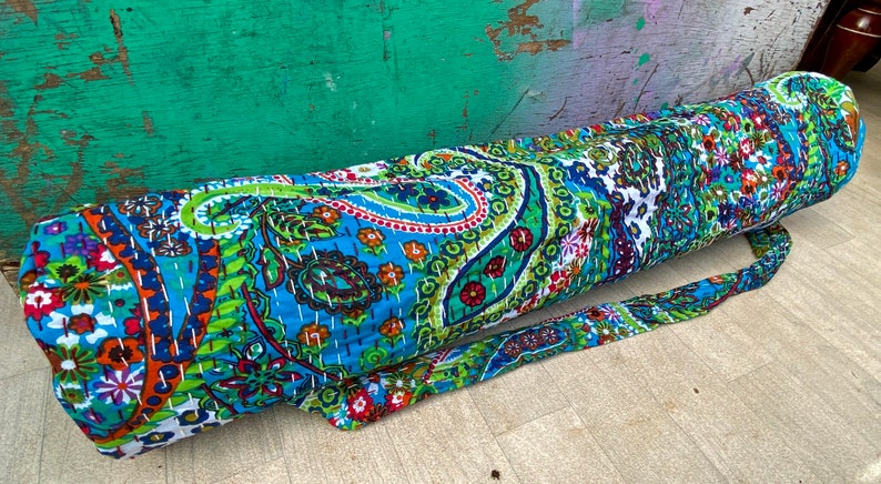 May include: A patterned yoga mat bag in shades of blue, green, orange, and white. The bag features a paisley and floral design and has a long strap for carrying. The bag is made of quilted fabric and is cylindrical in shape.