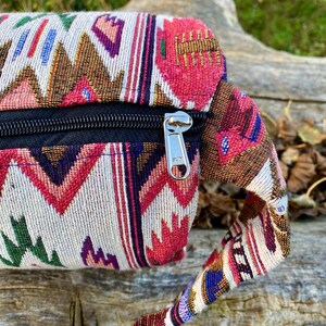 Pu&ograve; includere: Primo piano di un colorato marsupio intrecciato con cerniera nera. La borsa presenta un motivo geometrico nelle tonalit&agrave; del rosso, rosa, marrone, verde e blu su sfondo crema. Anche il cinturino &egrave; intrecciato con colori simili.