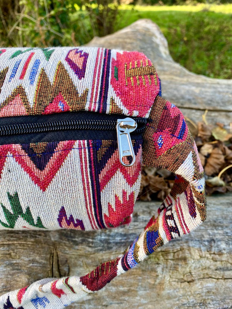 Pu&ograve; includere: Primo piano di un colorato marsupio intrecciato con cerniera nera. La borsa presenta un motivo geometrico nelle tonalit&agrave; del rosso, rosa, marrone, verde e blu su sfondo crema. Anche il cinturino &egrave; intrecciato con colori simili.
