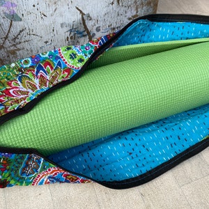 May include: A green yoga mat rolled up inside a colourful, patterned yoga bag. The bag features a blue paisley design with red, green, and yellow accents. The bag has a black zip and a black strap to secure the mat.