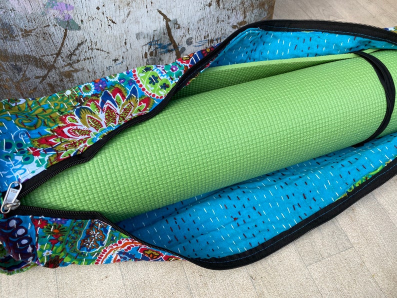 May include: A green yoga mat rolled up inside a colourful, patterned yoga bag. The bag features a blue paisley design with red, green, and yellow accents. The bag has a black zip and a black strap to secure the mat.