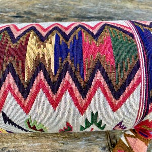 Pu&ograve; includere: Un rotolo di tessuto colorato a fantasia con un motivo a zig-zag. Il tessuto presenta un motivo ripetuto di zig-zag rossi, rosa, blu, verdi e oro su sfondo crema. Il rotolo &egrave; probabilmente un cuscino o un bolster decorativo.