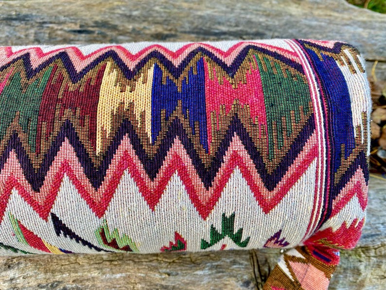 Pu&ograve; includere: Un rotolo di tessuto colorato a fantasia con un motivo a zig-zag. Il tessuto presenta un motivo ripetuto di zig-zag rossi, rosa, blu, verdi e oro su sfondo crema. Il rotolo &egrave; probabilmente un cuscino o un bolster decorativo.