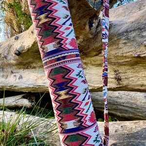 Pu&ograve; includere: Una colorata borsa per tappetino da yoga con un motivo a zigzag rosso, verde, blu e oro su sfondo bianco. La borsa ha una tracolla abbinata ed &egrave; esposta all'aperto, appoggiata a un tronco.