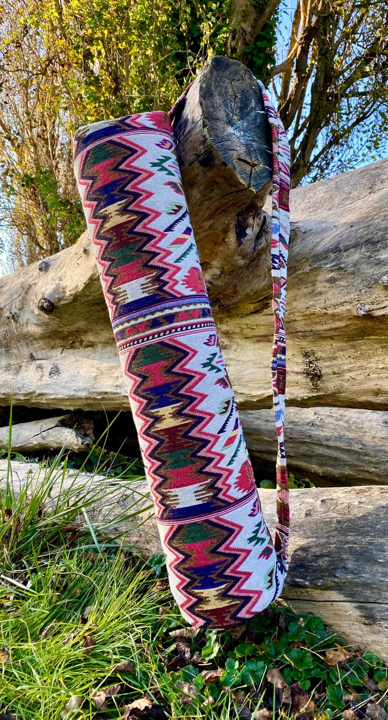 Pu&ograve; includere: Una colorata borsa per tappetino da yoga con un motivo a zigzag rosso, verde, blu e oro su sfondo bianco. La borsa ha una tracolla abbinata ed &egrave; esposta all'aperto, appoggiata a un tronco.