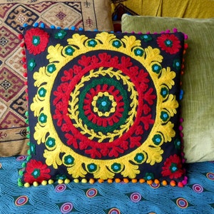 Puede incluir: Coj&iacute;n decorativo cuadrado con fondo azul oscuro y dise&ntilde;o central bordado en rojo, amarillo y verde. El dise&ntilde;o est&aacute; rodeado por un borde amarillo y adornado con pompones coloridos. El coj&iacute;n est&aacute; colocado sobre una superficie estampada.