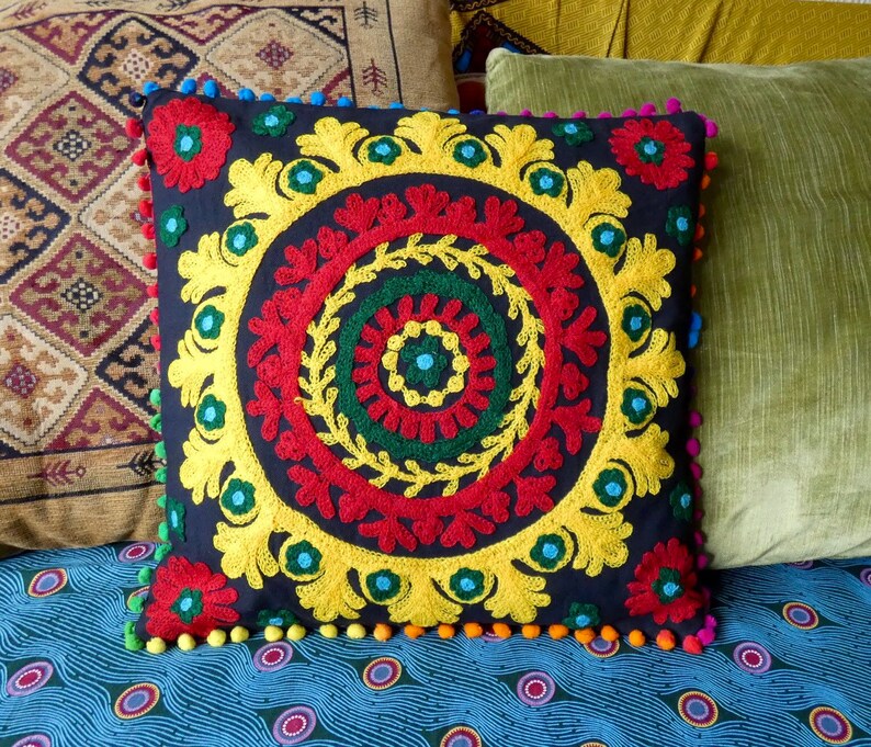 Puede incluir: Coj&iacute;n decorativo cuadrado con fondo azul oscuro y dise&ntilde;o central bordado en rojo, amarillo y verde. El dise&ntilde;o est&aacute; rodeado por un borde amarillo y adornado con pompones coloridos. El coj&iacute;n est&aacute; colocado sobre una superficie estampada.