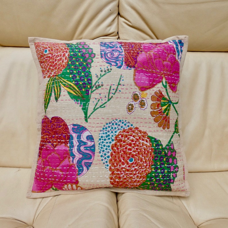 Puede incluir: Una almohada cuadrada con fondo blanco y un estampado floral colorido. La almohada presenta flores rosas, naranjas, verdes y azules con intrincados detalles de costura.