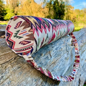Pu&ograve; includere: Un bolster da yoga cilindrico con una fodera in tessuto colorato e fantasia. Il design presenta un mix di motivi geometrici e tribali nei toni del rosso, blu, verde e marrone. Ha un manico in tessuto abbinato.