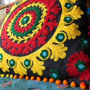 Puede incluir: Un coj&iacute;n cuadrado negro con un vibrante dise&ntilde;o floral bordado en rojo, amarillo, verde y azul. El coj&iacute;n tiene un borde de peque&ntilde;os pompones naranjas y es un art&iacute;culo de decoraci&oacute;n para el hogar.
