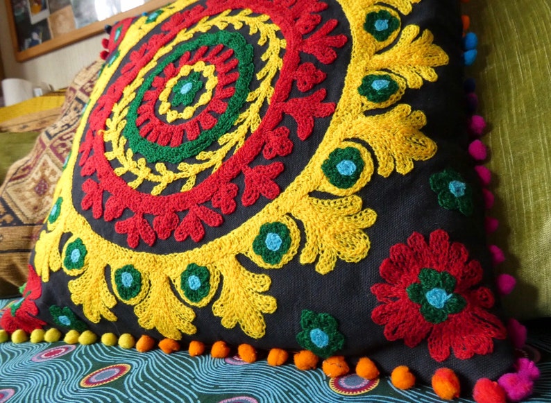 Puede incluir: Un coj&iacute;n cuadrado negro con un vibrante dise&ntilde;o floral bordado en rojo, amarillo, verde y azul. El coj&iacute;n tiene un borde de peque&ntilde;os pompones naranjas y es un art&iacute;culo de decoraci&oacute;n para el hogar.