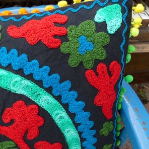 May include: A black pillow with colorful embroidered designs. The pillow has a fringe border with yellow pom-poms.