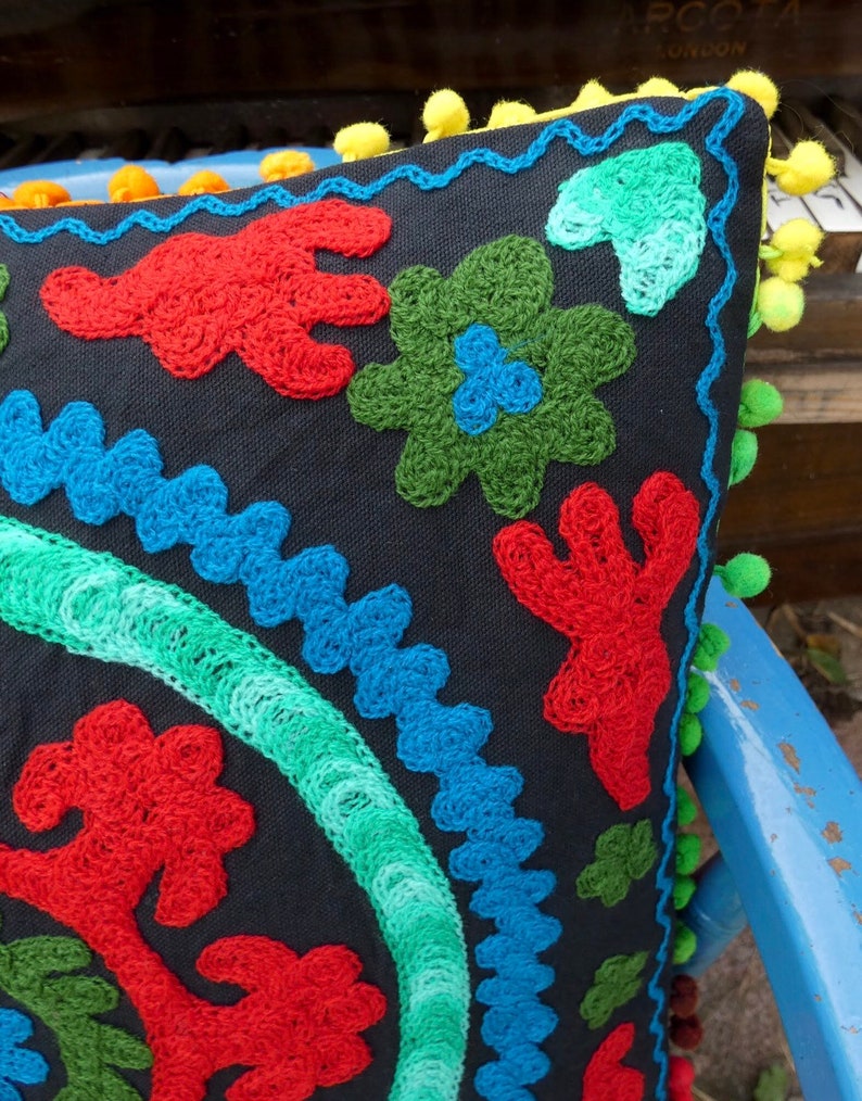 May include: A black pillow with colorful embroidered designs. The pillow has a fringe border with yellow pom-poms.