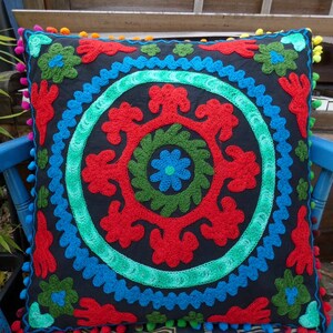 May include: A black square throw pillow with a colorful embroidered design. The design features a circle with red, green, and blue floral patterns.