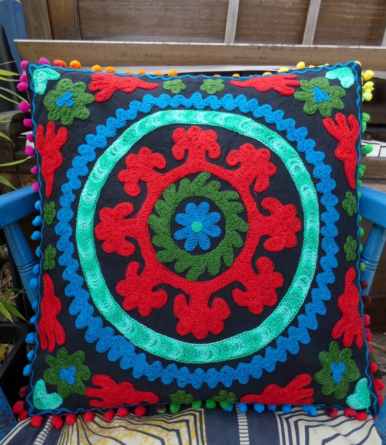 May include: A black square throw pillow with a colorful embroidered design. The design features a circle with red, green, and blue floral patterns.