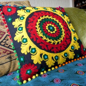 Puede incluir: Coj&iacute;n decorativo cuadrado con fondo azul oscuro y dise&ntilde;o floral bordado colorido. El bordado presenta detalles en rojo, amarillo, verde y azul. El coj&iacute;n est&aacute; adornado con pompones coloridos.