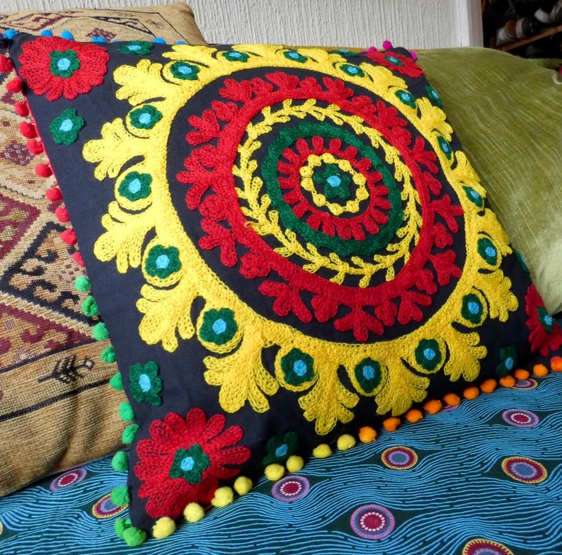 Puede incluir: Coj&iacute;n decorativo cuadrado con fondo azul oscuro y dise&ntilde;o floral bordado colorido. El bordado presenta detalles en rojo, amarillo, verde y azul. El coj&iacute;n est&aacute; adornado con pompones coloridos.