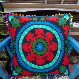 May include: A black throw pillow with a colorful embroidered design. The design features a circle with red, green, and blue floral patterns. The pillow has a pom-pom trim.