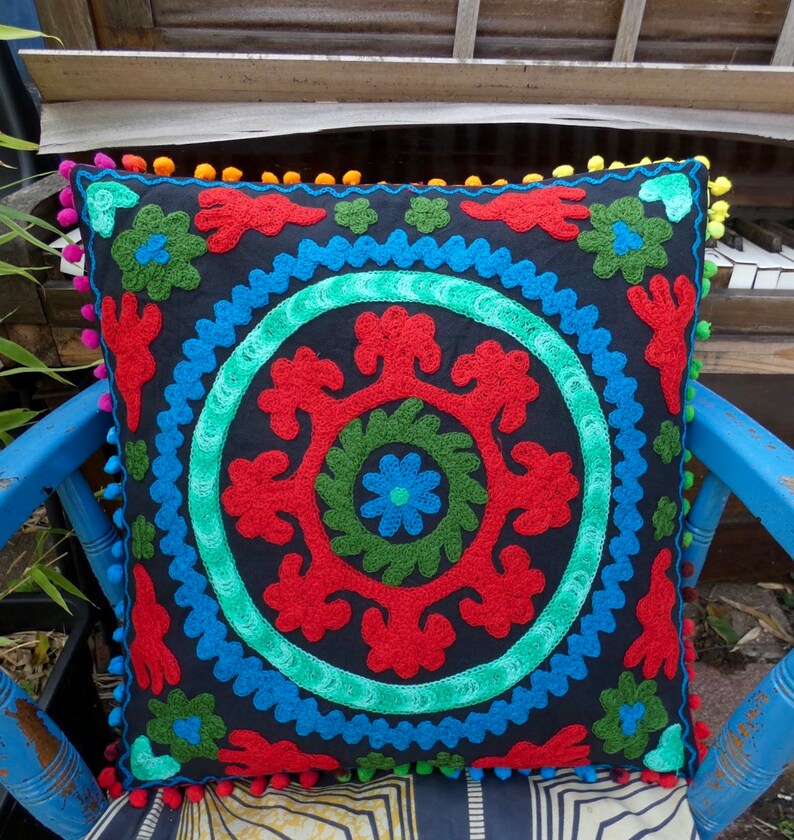 May include: A black throw pillow with a colorful embroidered design. The design features a circle with red, green, and blue floral patterns. The pillow has a pom-pom trim.
