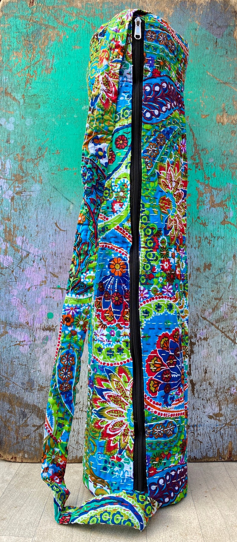 May include: A patterned yoga mat bag in blue, green, red, and orange. The bag features a black zip and a matching strap. The bag is made of a quilted fabric with a floral design. The bag is designed to carry a yoga mat.