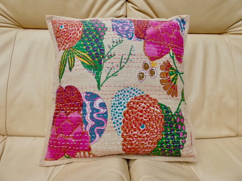 Puede incluir: Una almohada cuadrada con fondo blanco y un estampado floral colorido. La almohada presenta flores rosas, naranjas, verdes y azules con intrincados detalles de costura.
