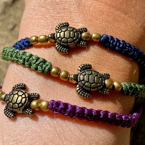 Op de afbeelding: Drie armbanden met gouden schildpadbedels. De armbanden zijn gemaakt van gevlochten koord in paars, groen en blauw.