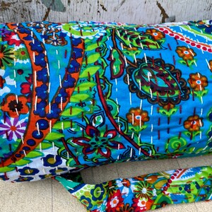 May include: A rolled fabric item with a blue background and colourful floral and paisley patterns. The fabric is stitched with white thread. A matching strap is attached to the item.