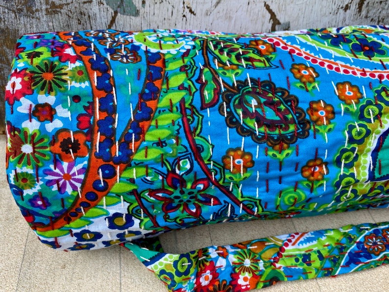 May include: A rolled fabric item with a blue background and colourful floral and paisley patterns. The fabric is stitched with white thread. A matching strap is attached to the item.
