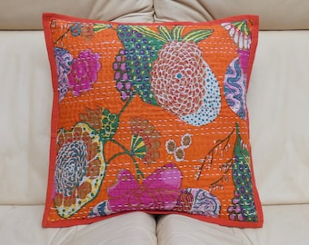 May include: Square orange pillow with a floral design in pink, green, and blue. The pillow has a red border and white stitching. The pillow is likely made of fabric.
