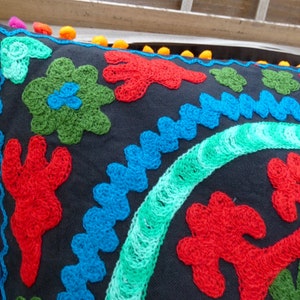 May include: A black pillow with colorful embroidered flowers and a blue and green border. The pillow has a pom-pom trim.