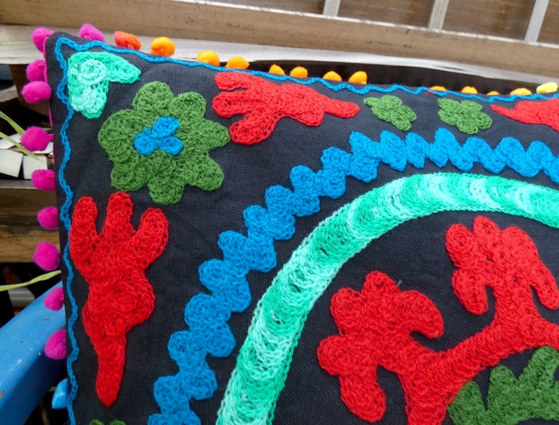 May include: A black pillow with colorful embroidered flowers and a blue and green border. The pillow has a pom-pom trim.