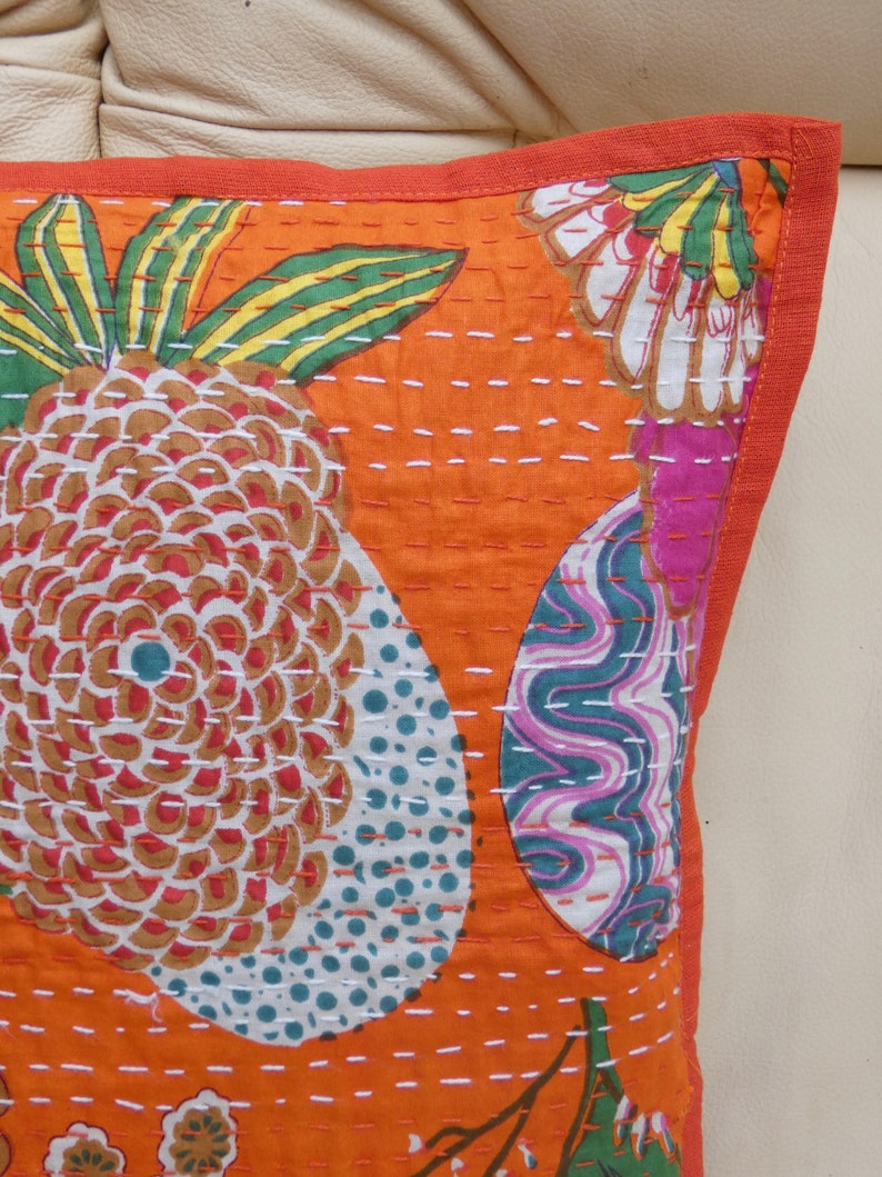 Peut inclure: Gros plan sur un coussin d&eacute;coratif orange avec un motif d'ananas et floral. Le coussin pr&eacute;sente un fond orange vif avec des coutures blanches et une bordure rouge. L'ananas a un motif marron, blanc et rouge.