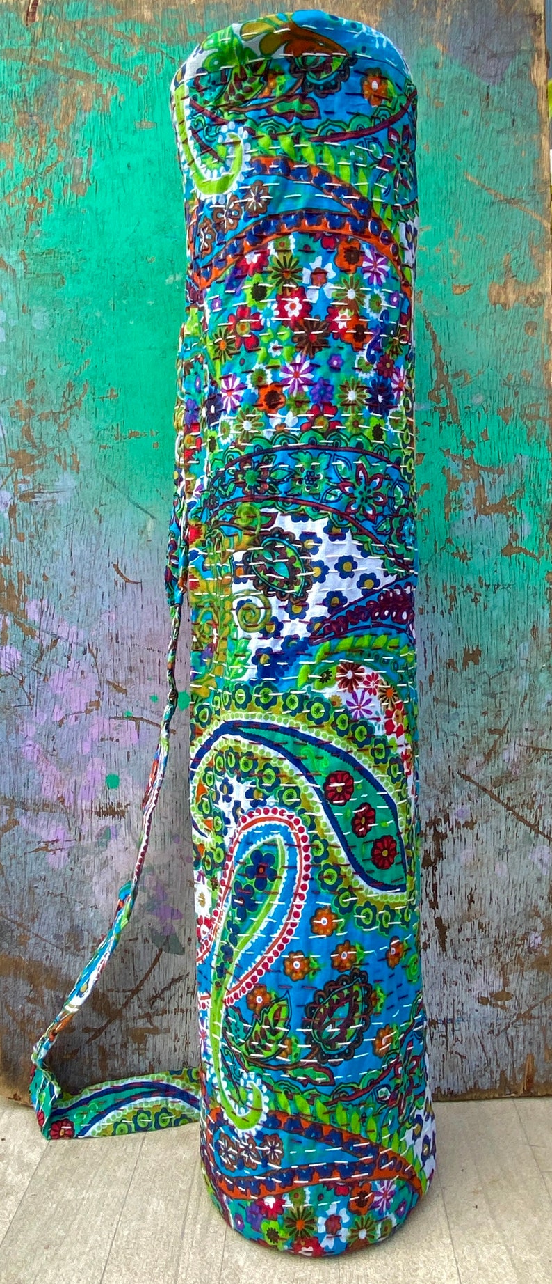 May include: A cylindrical yoga mat bag with a vibrant paisley design in blue, green, red, and white. The bag features a matching strap and is constructed from stitched fabric.