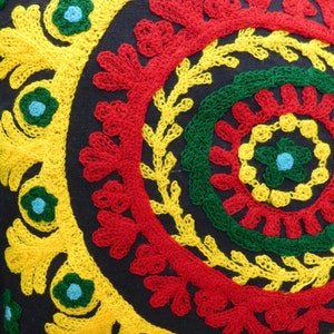 Puede incluir: Primer plano de un coj&iacute;n decorativo con fondo azul oscuro. El coj&iacute;n presenta un dise&ntilde;o bordado vibrante con c&iacute;rculos conc&eacute;ntricos en rojo, amarillo y verde. Peque&ntilde;os acentos florales azules a&ntilde;aden toques de color al dise&ntilde;o.