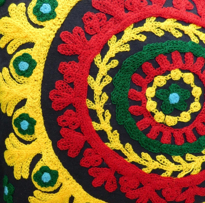 Puede incluir: Primer plano de un coj&iacute;n decorativo con fondo azul oscuro. El coj&iacute;n presenta un dise&ntilde;o bordado vibrante con c&iacute;rculos conc&eacute;ntricos en rojo, amarillo y verde. Peque&ntilde;os acentos florales azules a&ntilde;aden toques de color al dise&ntilde;o.