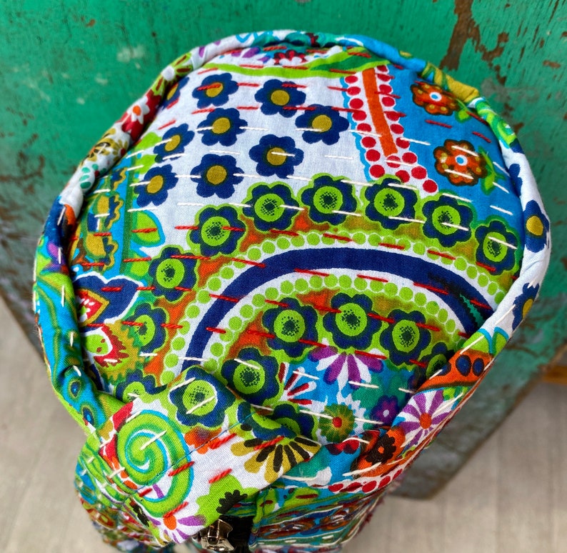 May include: A circular bag with a zip. The bag is decorated with a colourful floral and paisley pattern in shades of blue, green, orange, and white. It is made from quilted fabric and has a stitched edge.