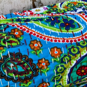May include: A colourful, hand-stitched textile with a paisley and floral design. The fabric has a bright blue background with green, orange, and red accents. White stitching outlines the patterns, creating a textured appearance. Suitable for home decor.