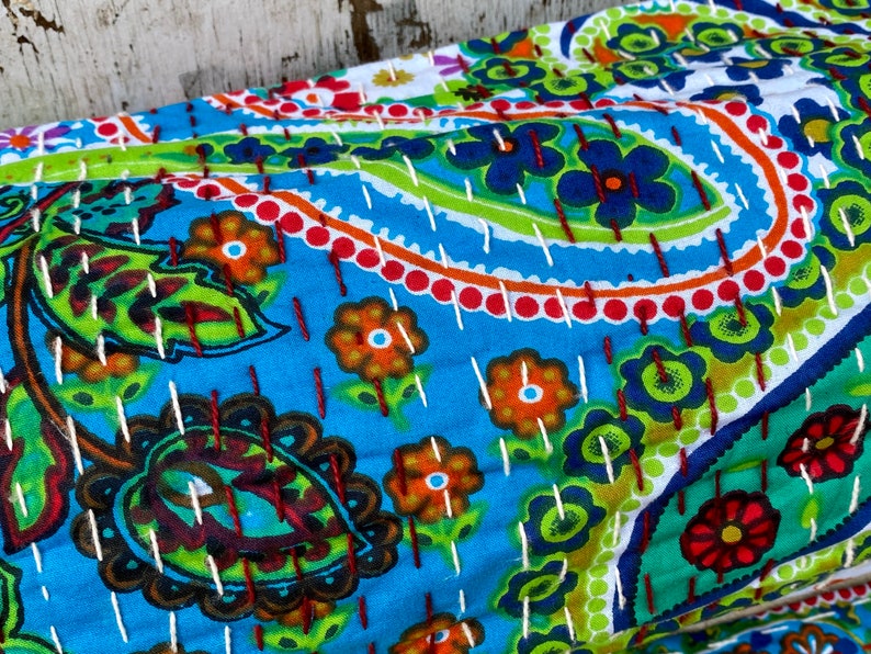 May include: A colourful, hand-stitched textile with a paisley and floral design. The fabric has a bright blue background with green, orange, and red accents. White stitching outlines the patterns, creating a textured appearance. Suitable for home decor.