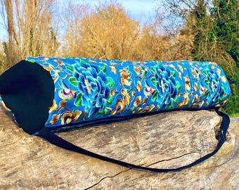 Pode incluir: Uma bolsa cilíndrica para tapete de ioga com um vibrante padrão floral azul. A bolsa apresenta grandes flores azuis estilizadas com detalhes dourados e verdes. Possui tampas pretas e uma alça de ombro preta ajustável.